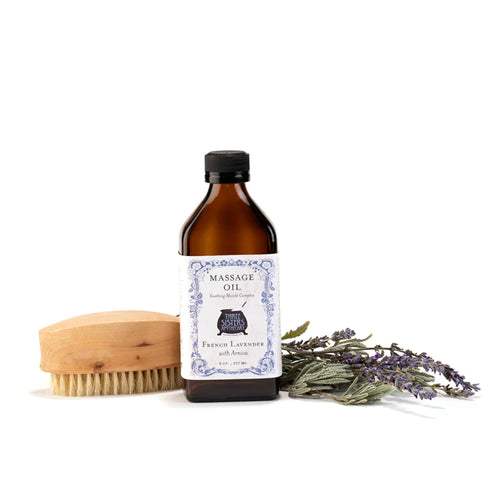 A bottle of Three Sisters Apothecary French Lavender Massage Oil with essential fatty acids features a vintage-style label beside a wooden brush and a bunch of lavender on a white background.