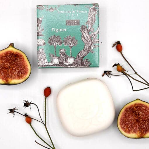A flat lay composition with a Senteurs De France fig-scented soap, a book titled "Senteurs de France," half-cut figs, and delicate twigs with small red berries on a white background.