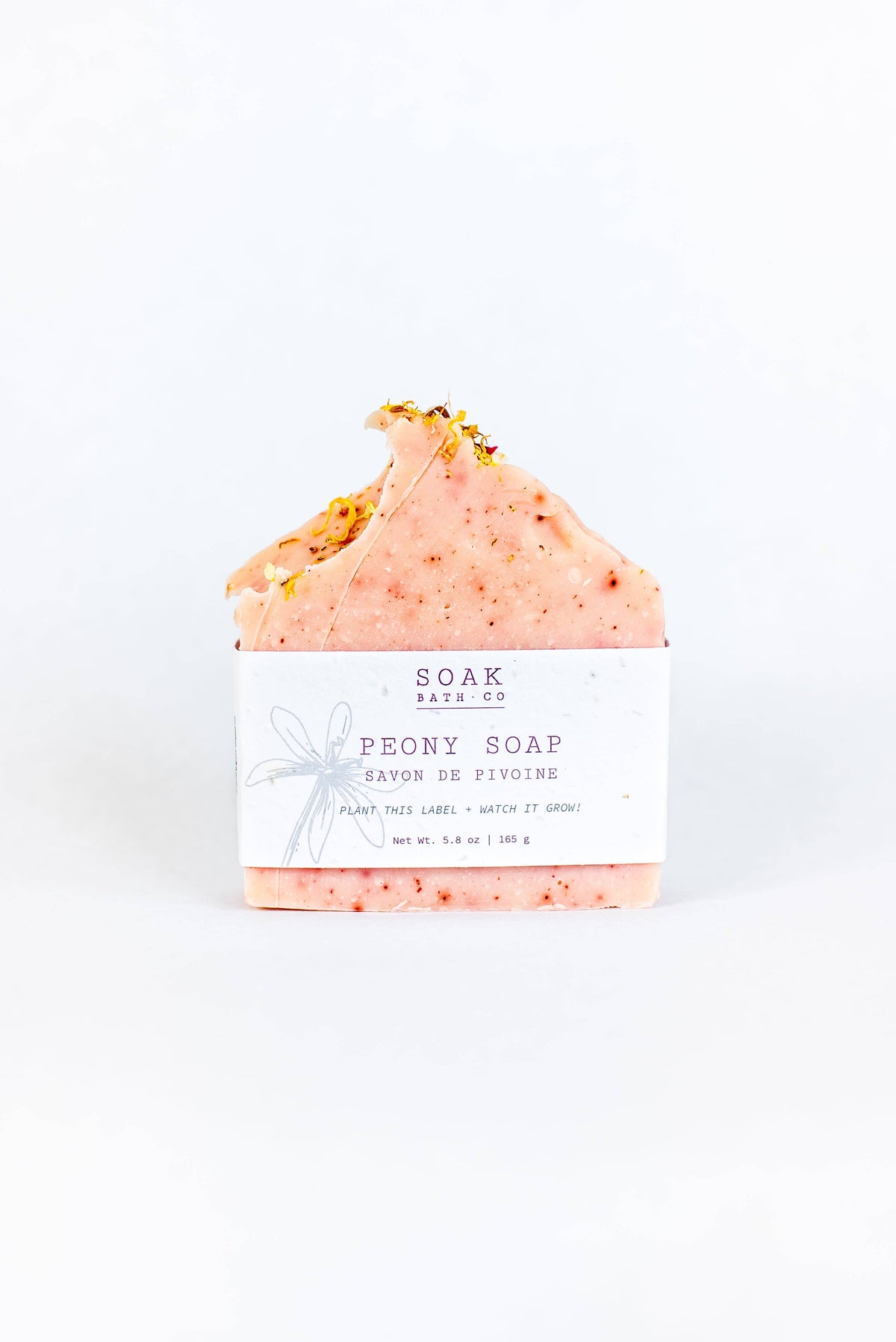 A SOAK Bath Co. - Peony Soap Bar with gold flakes on top, wrapped in plantable seed paper, against a white background. The label features a drawn bow and text describing the product.