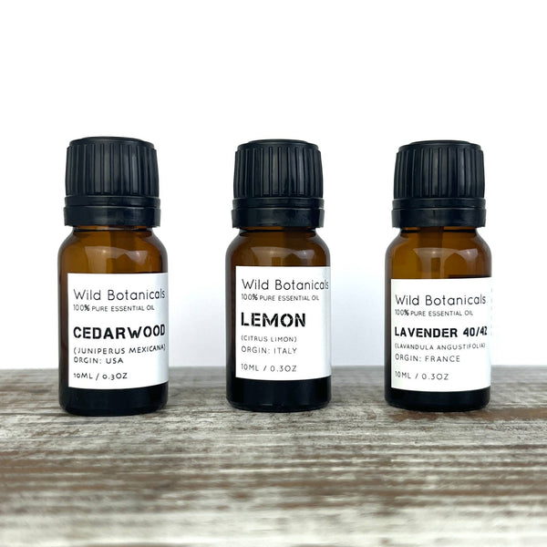 Three amber-colored glass bottles of Wild Botanicals essential oils are arranged side by side on a wooden surface. The labels read "Wild Botanicals Citronella Essential Oil" (origin: USA), "Lemon" (origin: Italy), and "Lavender 40/42" (origin: France). Renowned for flu protection and cold relief, each bottle is labeled as 10mL (0.3 oz).