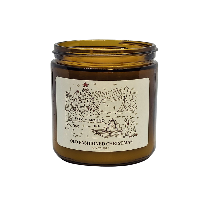 A Fox + Hound odor eliminator in a small amber glass jar labeled "Old Fashioned Christmas" featuring a wintery scene with a cabin, trees, and sled, under the brand "Fox + Hound.