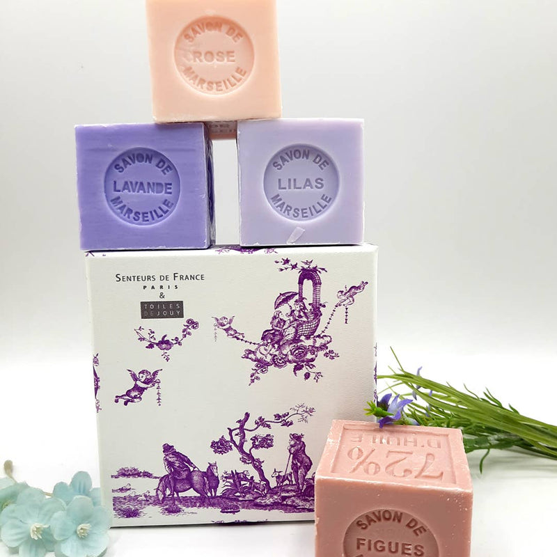 Four handmade Senteurs De France Coffret Lavender, Lilac, Fig and Rose Soaps stacked beside a vintage-style printed box depicting rural scenes, on a white background with greenery.