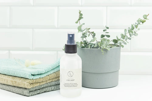 A white spray bottle labeled "Lizush Eucalyptus Mist Spray" sits beside a stack of folded towels and a gray pot containing a green eucalyptus plant, against a white tiled background.