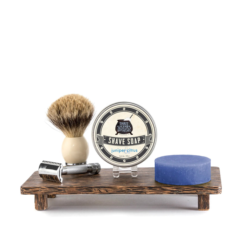 A shaving kit featuring a badger hair brush, a safety razor, a tin of Three Sisters Apothecary Shave Soap - Juniper & Citrus enriched with essential oils, and a blue bar of soap, all set on a small wooden stand.