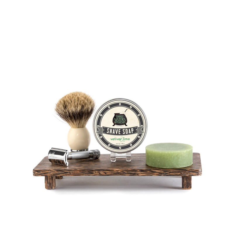 Shaving kit on a wooden stand featuring a safety razor, a shaving brush, Three Sisters Apothecary Shave Soap - Vetiver & Lime with natural ingredients, and a green bar of soap, against a