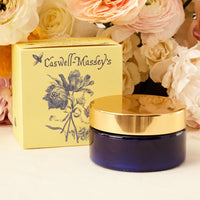 A Caswell Massey Elixir of Love Body Cream jar next to its packaging box, adorned with a floral design, placed against a backdrop of soft-focus flowers.