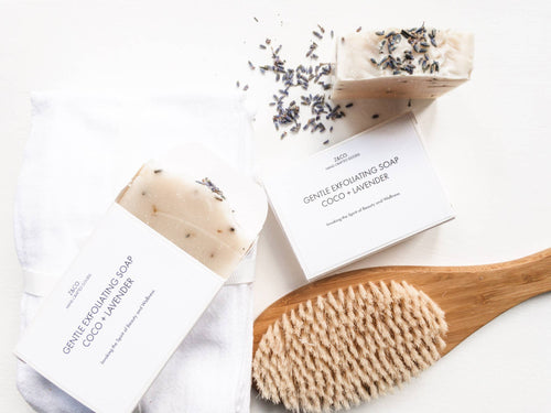A flat lay of Z&Co. CoCo + Lavender Soap bars with dried lavender on top, accompanied by their packaging and a wooden body brush, all arranged on a light, textured surface.