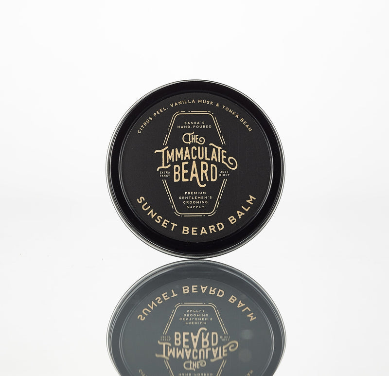 A round tin of "The Immaculate Beard Dusk Beard Balm-Made in Texas" on a reflective surface, displaying the logo and product name in stylized text. The label colors are black and gold.