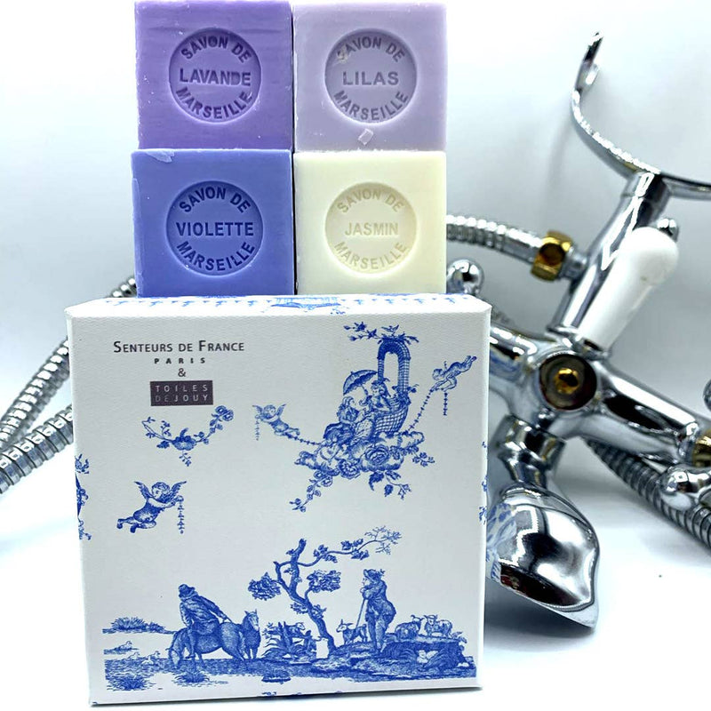 A stack of Senteurs De France Coffret Lavender, Violet, Lilac, Jasmine Cube Soaps beside a silver chrome faucet on a white background. The soap packaging features intricate Toile de Jouy designs.
