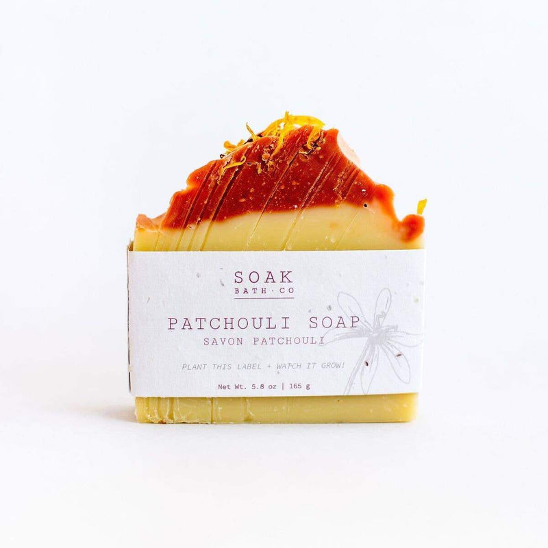 A bar of SOAK Bath Co. - Patchouli Soap with orange drizzle on top, presented on a white background. The soap is wrapped with a zero-waste label that includes the text "soak" and