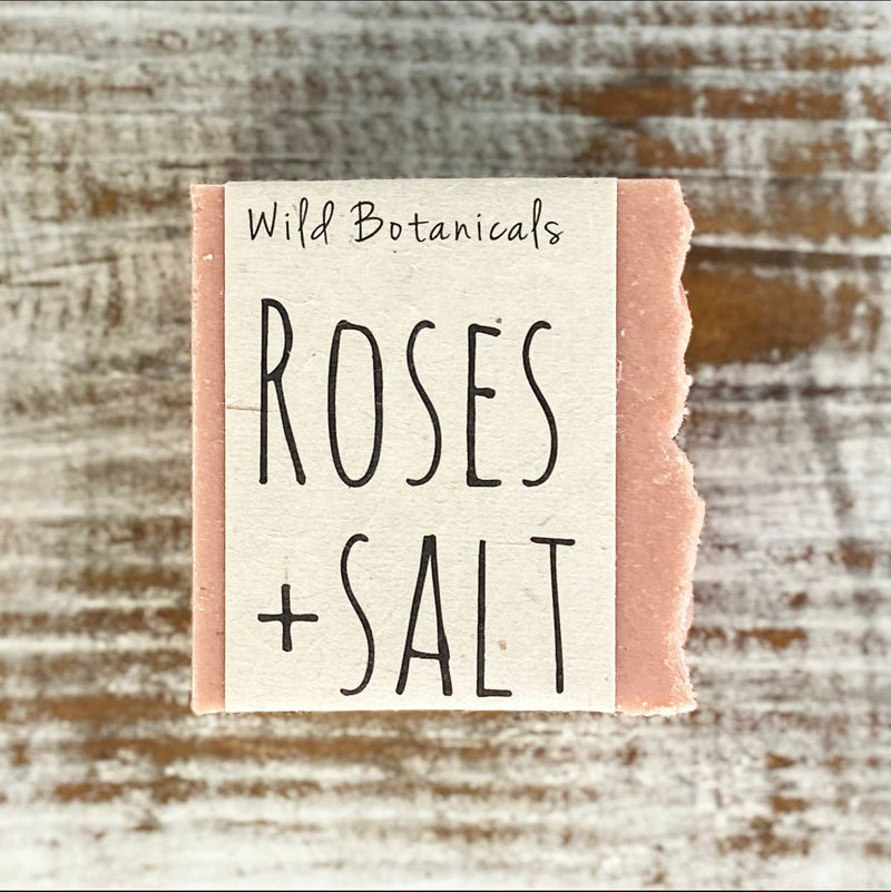 A handmade soap labeled "Wild Botanicals Roses and Himalayan Salt Soap" rests on a rustic wooden surface. The soap has a rough, textured edge and soft pink color.