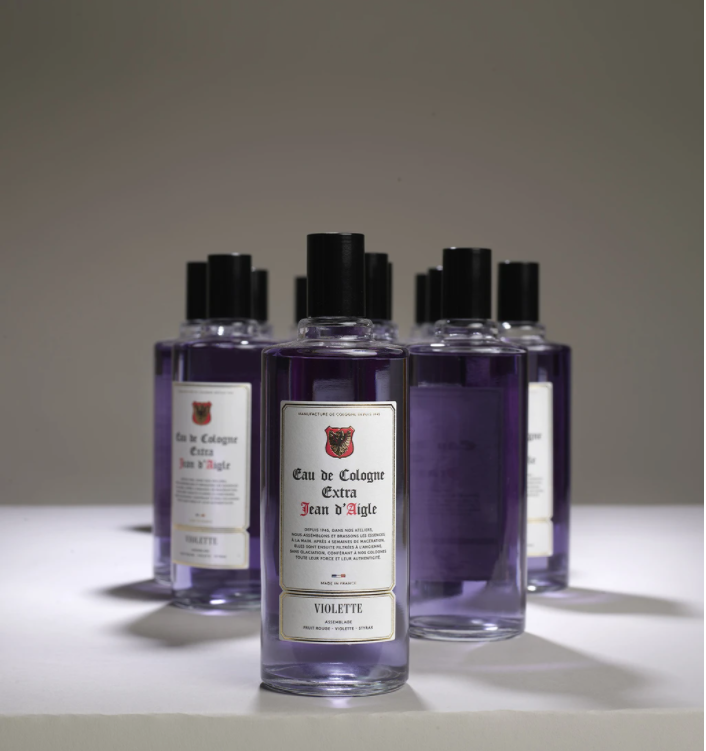 Five bottles of violet-colored cologne labeled "Jean d'Aigle Violette Eau de Cologne" arranged in a staggered line, with the front bottle in sharp focus and the rest slightly blurred.
