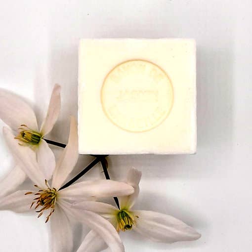 A square, handmade Senteurs De France Marseille Jasmine Cube soap with embossed text next to delicate white flowers on a plain white background.