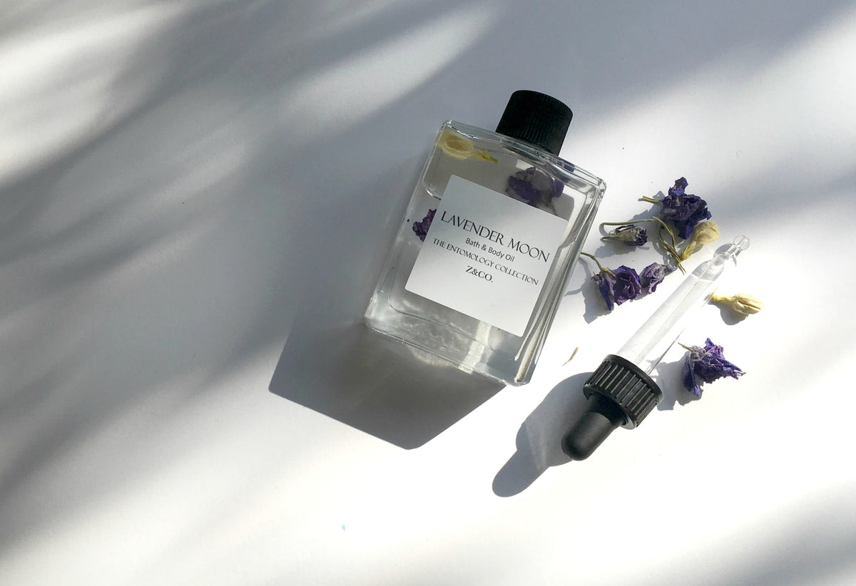 A bottle of Z&Co. Lavender Moon Bath & Body Oil and a dropper, surrounded by dried purple flowers, placed under sunlight casting striped shadows, on a white surface.