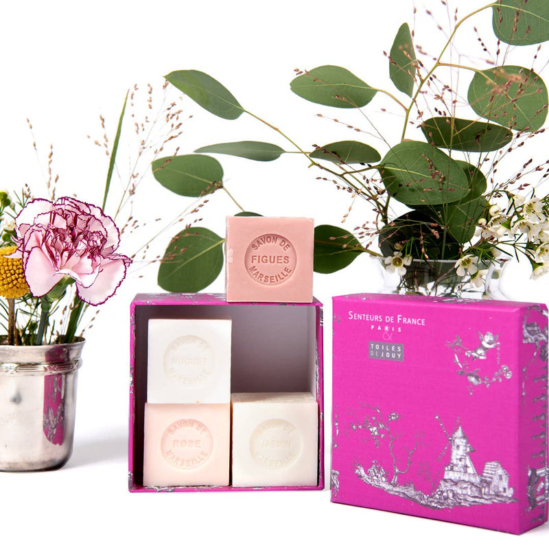 An elegant arrangement of Senteurs De France Coffret of Rose, Lily of the Valley, Fig & Jasmine Cube Soaps in an open pink box alongside a bouquet of wildflowers and greenery in a glass jar, isolated on a white background.