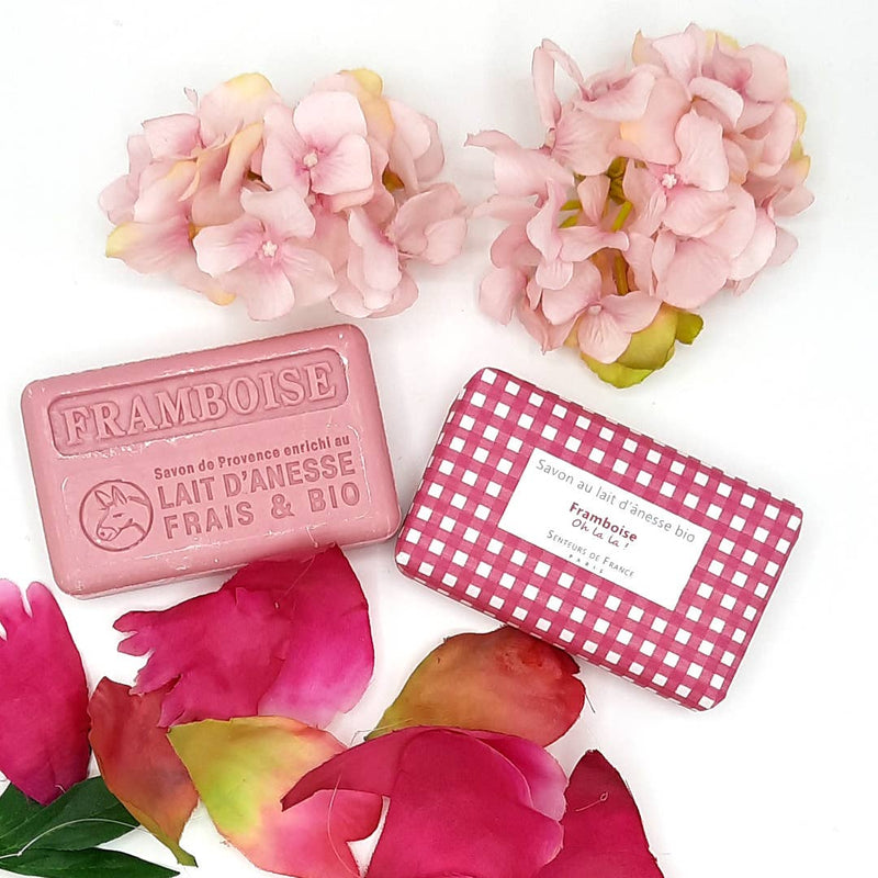 Two bars of Senteurs De France Vichy Raspberry Soap, surrounded by pink petals and pale pink flowers on a white background. One soap is labeled "Vichy Raspberry Soap" and the other "organic.