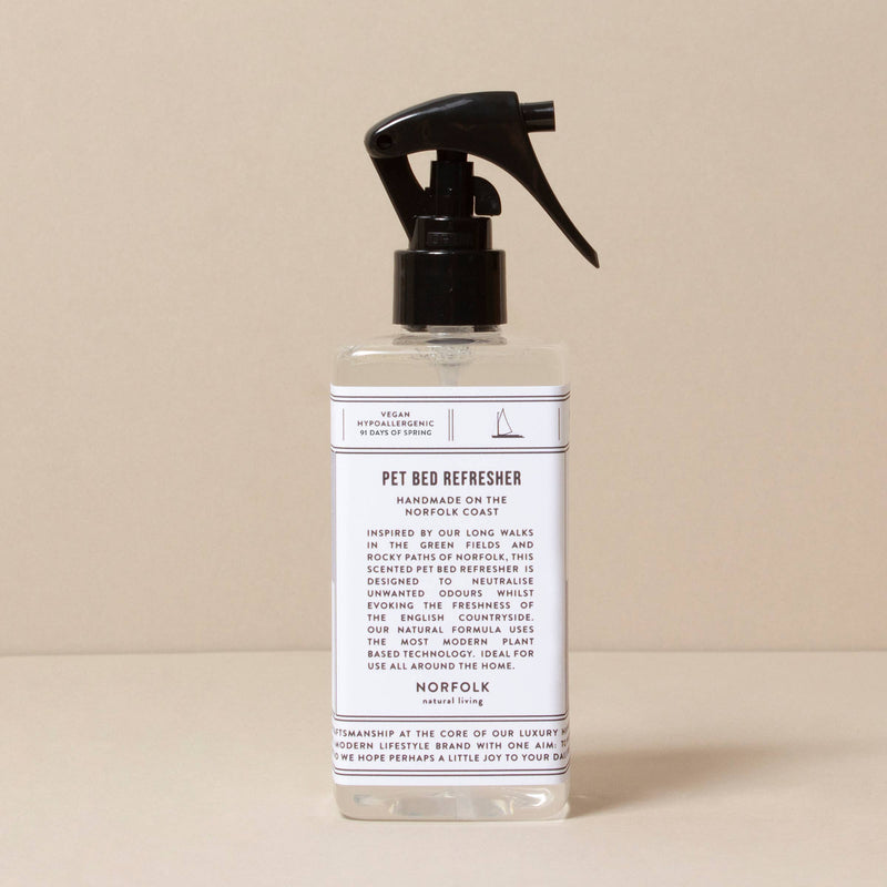 A bottle of "Norfolk Natural Living Spring 91 Pet Bed Refresher" with a spray nozzle, featuring a beige background. The label lists vegan, natural anti-bacterial ingredients and instructions for use to keep pet beds fresh and clean.