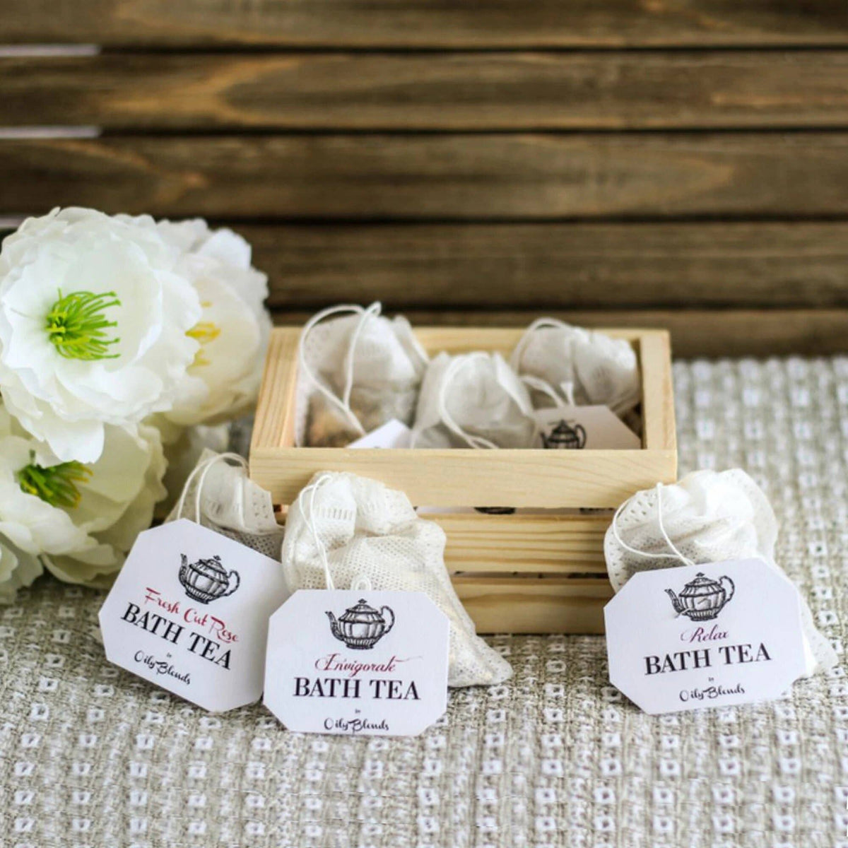 A wooden crate holding small pouches labeled "Hampton Court herbal bath tea," alongside large white flowers on a textured surface. The setting conveys a serene, spa-like atmosphere.