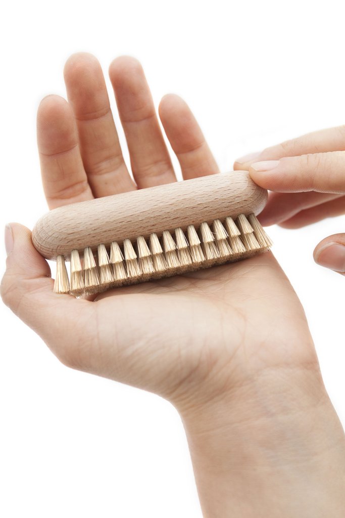 A close-up of a hand holding a small Andrée Jardin Tradition Beech Wood Nail Brush with stiff bristles, against a white background. The fingers of the other hand are touching the bristles.