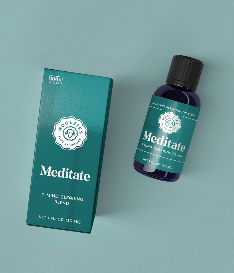 A box and dark glass bottle labeled "Woolzies Meditate Blend," a mind-clearing essential oil blend aimed at promoting restful sleep. The packaging features teal and dark blue colors with a floral emblem.