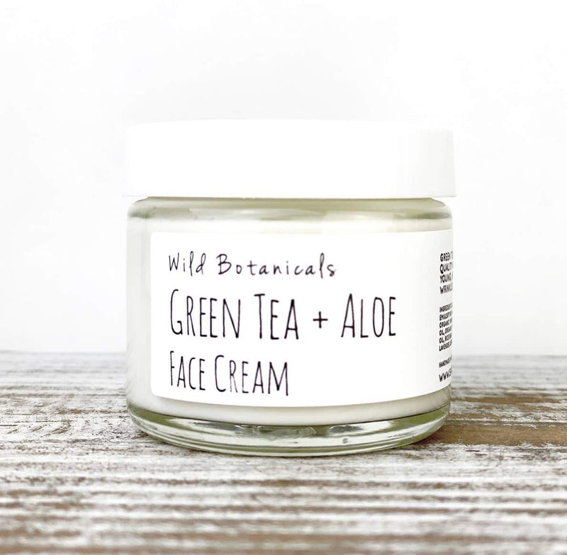 A clear glass jar of Wild Botanicals Green Tea and Aloe Face Cream sits on a wooden surface against a white background.