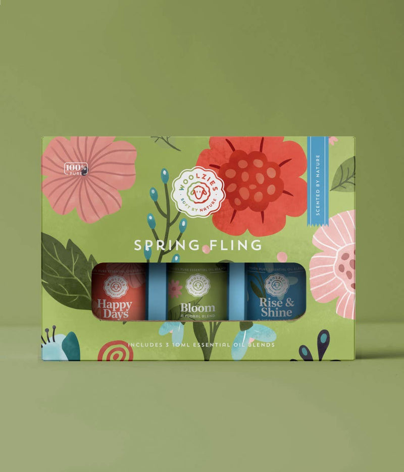 A colorful box labeled "Woolzies - The Spring Fling Collection" featuring floral designs and containing three essential oil blends labeled "happy days," "bloom," and "rise & shine" against a green