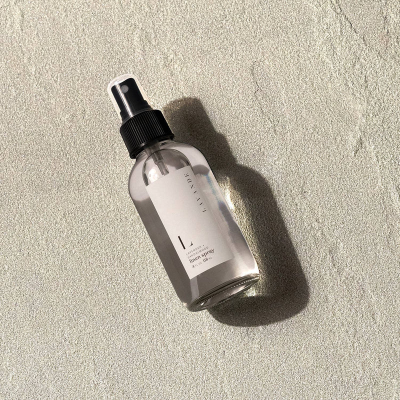 A clear glass spray bottle labeled "Lavande - Lavender & Sandalwood Linen Spray 4oz" rests on a textured concrete surface in sunlight, casting a soft shadow.