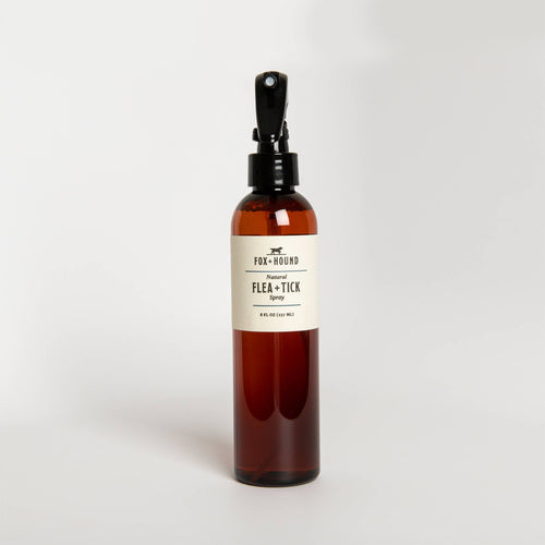 A brown glass spray bottle labeled "Fox + Hound - Natural Flea with Tick Spray For Dogs" on a white background. The bottle contains a reddish liquid and has a black spray nozzle.