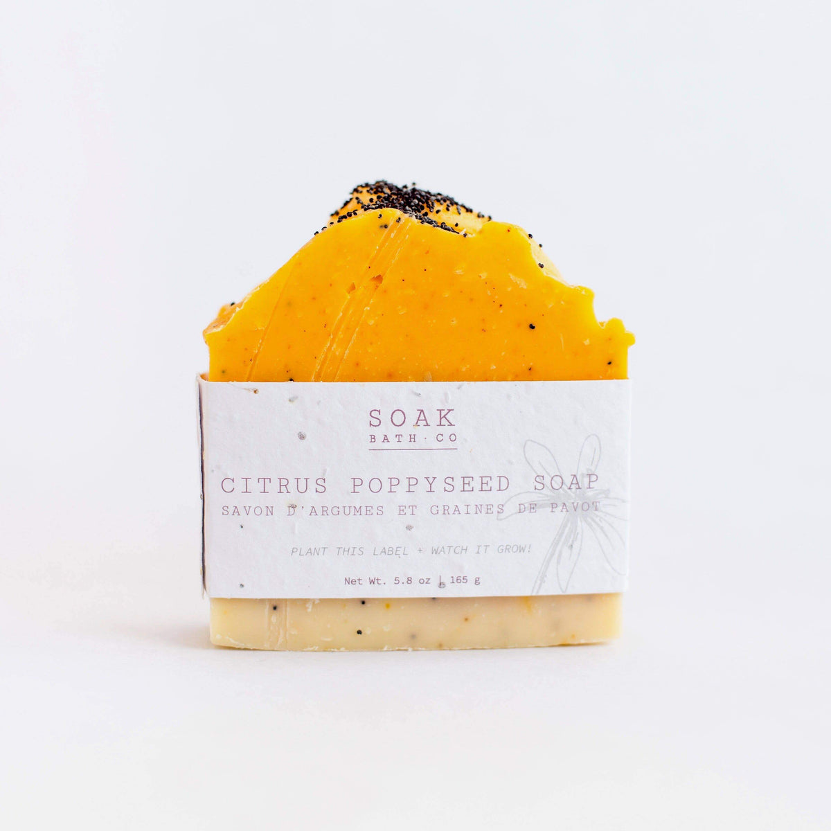 A bar of SOAK Bath Co. - Citrus Poppyseed Soap, wrapped in labeled paper that can be planted. This zero waste soap is topped with a yellow, textured surface sprinkled with black poppy seeds.