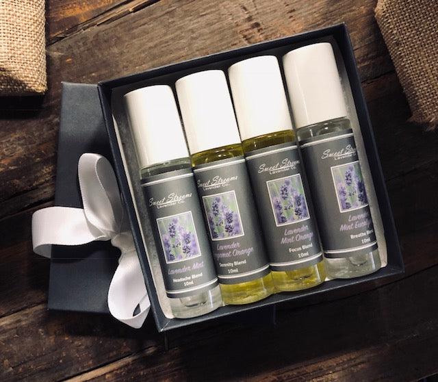 Four bottles of Sweet Streams Lavender Co. lavender mint headache blend oils in a dark blue gift box, adorned with a white ribbon, displayed on a rustic wooden surface.