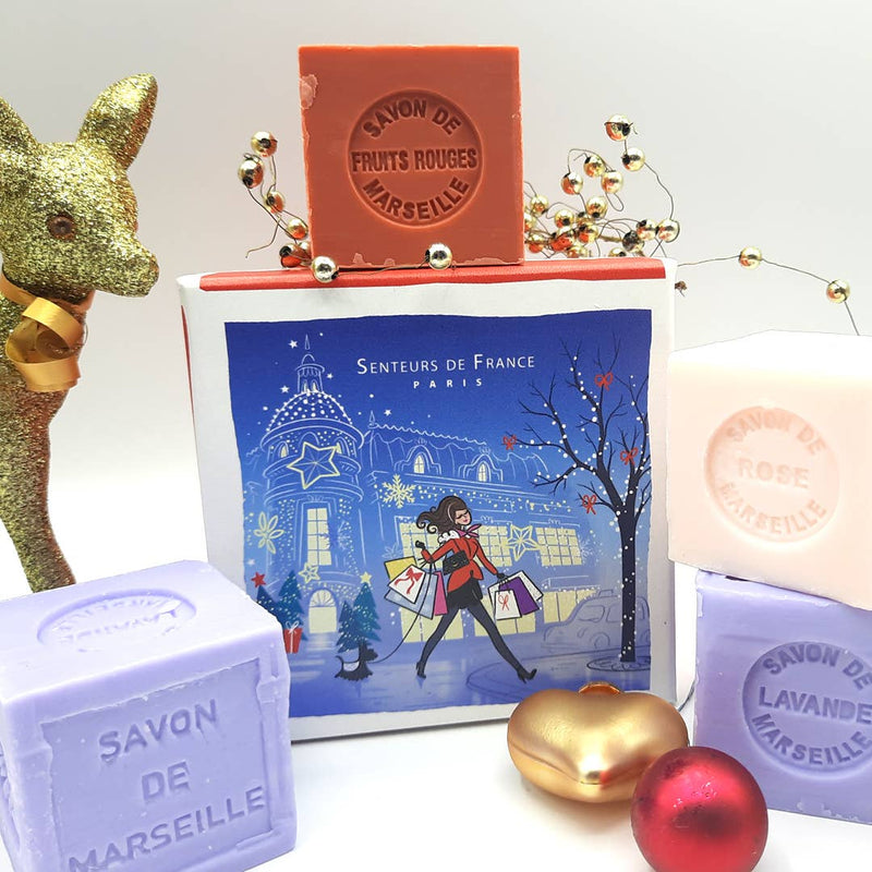 A festive display featuring various Senteurs De France natural soaps labeled "savon de marseille", with a holiday-themed box depicting a shopper in Paris. Decor includes a gold reindeer and red baubles.