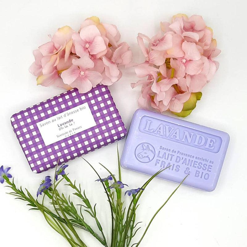 A purple gingham-patterned soap box and a Senteurs De France Vichy Lavender Soap bar marked "lavande" with flowers, including pink hydrangeas and blue lavender, arranged around on a white background.