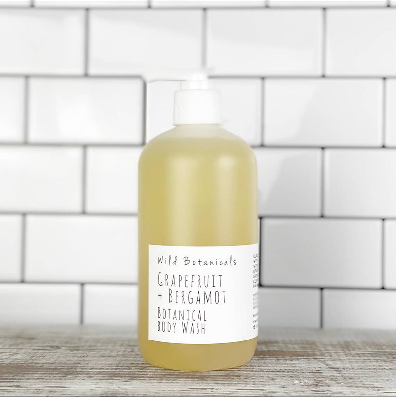 A large, transparent bottle of Wild Botanicals - Grapefruit & Bergamot Botanical Body Wash stands in front of a white brick wall. The bottle, featuring essential oils, has a white label with black text.