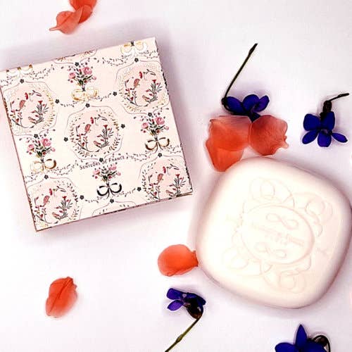A Senteurs De France Lavender Soap "Versailles" carved with a floral design beside a matching floral patterned box, surrounded by scattered petals and blue flowers on a white background.