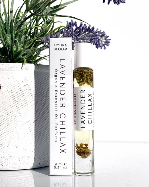 A clear roller bottle labeled "Hydra Bloom Beauty Lavender Chillax" with visible flowers inside, standing next to a Hydra Bloom box and a potted plant with lavender, all against a white background. This essential.