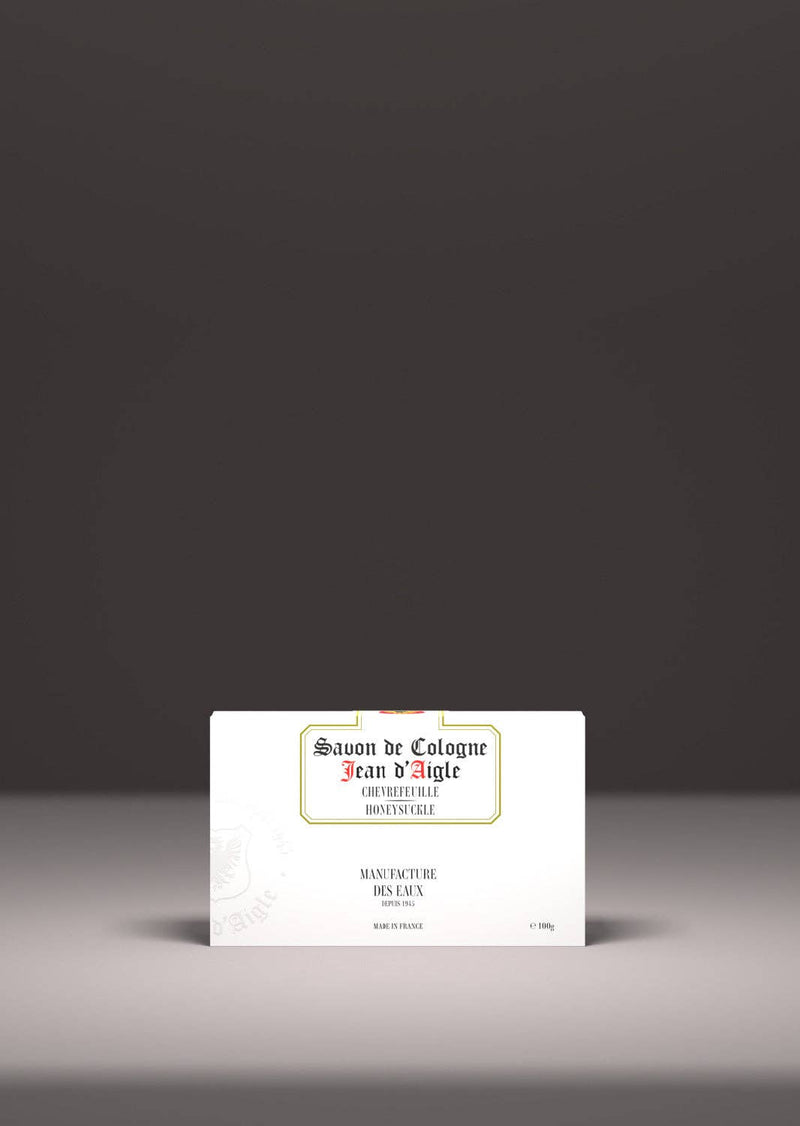 A vintage-style Jean d'Aigle Honeysuckle Soap label featuring elegant typography, displayed centered against a soft gray backdrop. The label has ornate text and decorative elements that evoke a floral aroma.