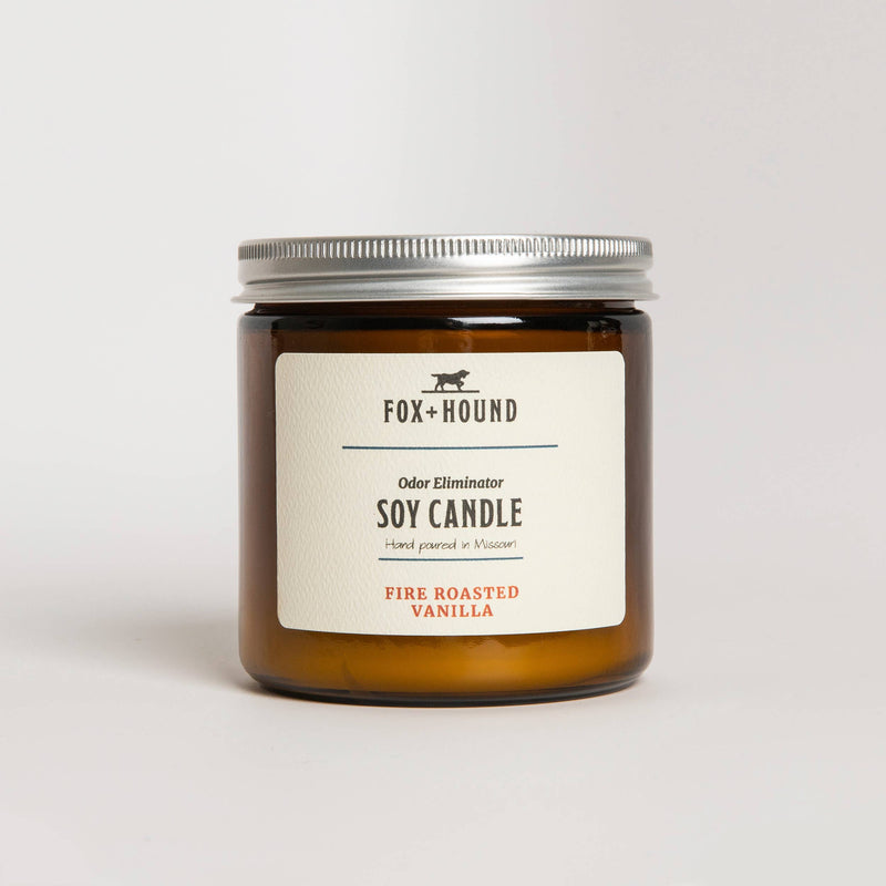 A clear glass jar candle labeled "Fox + Hound Odor Eliminator Soy Candle - Fire Roasted Vanilla" with a beige label and black cap, set against a white background, designed as an odor-eliminating soy candle