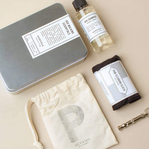 A neatly arranged Norfolk Natural Living Dog Walking Essentials Kit on a beige background, including a tin box, a bottle labeled "dr. shaving moroka," a dark cloth, a biodegradable bag with a letter.