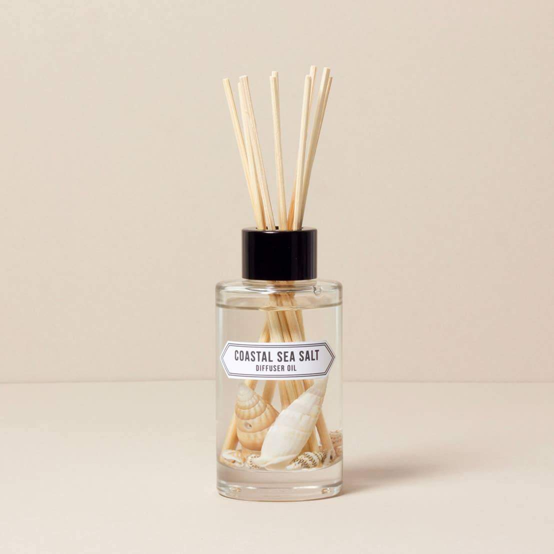A Norfolk Natural Living reed diffuser oil bottle labeled "Coastal - Sea Salt" with multiple sticks inserted, set against a plain beige background. The bottle features a design with seashells.