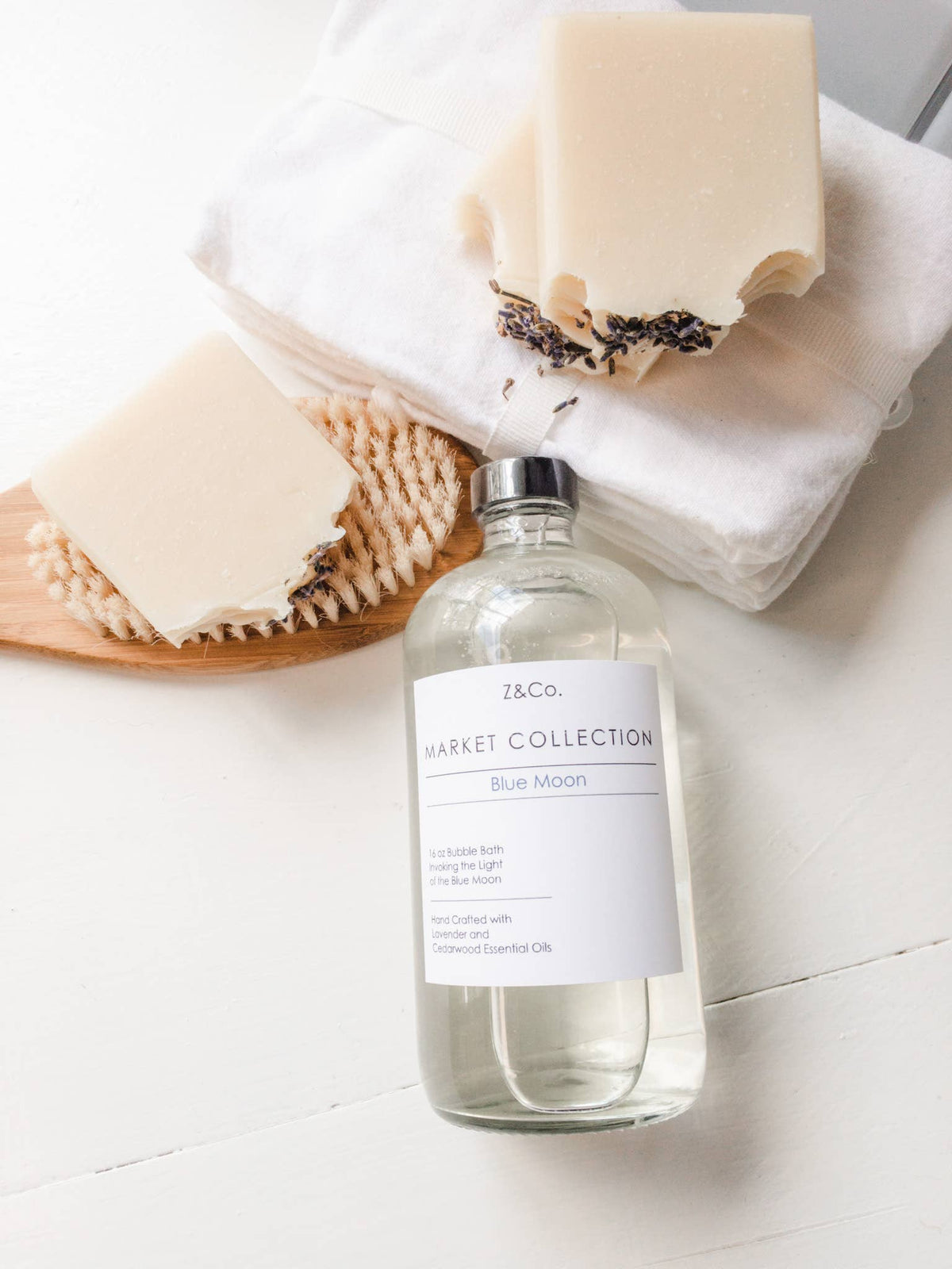 Two bars of soap with lavender essential oil on a wooden soap dish beside a white towel and a Z&Co. Market Collection Blue moon Bubble Bath in a clear bottle on a white surface.