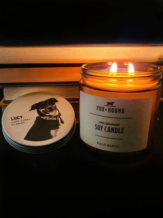 A Fox + Hound Rescue Dog Lucy Odor Eliminating Soy Candle Palo Santo in a glass jar with a label reading "rescue dog candles palo santo" next to its closed lid showing a picture of an emotional support dog named Lucy, illuminated by