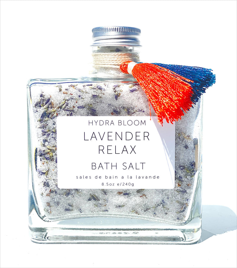 A square glass bottle filled with Hydra Bloom Beauty Lavender Relax Bath Salts, labeled "Hydra Bloom Beauty Lavender Relax Bath Salt," capped with a metal lid and decorated with a red and blue tassel against a white background. Contains