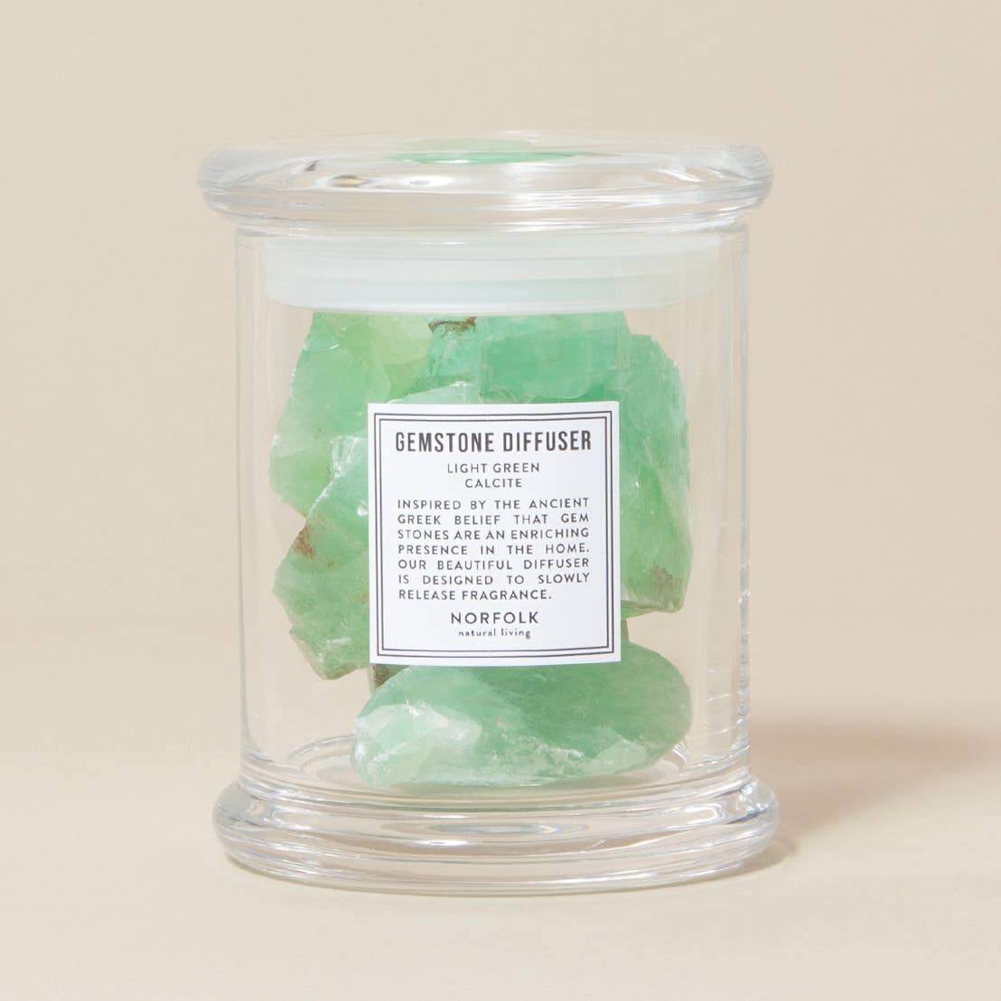 A clear jar filled with Norfolk Natural Living Coastal Gemstone Diffuser - Light Green Calcite gemstones, labeled "gemstone diffuser - calcite" detailing its use to infuse spaces with delicate, naturally releasing fragrance.