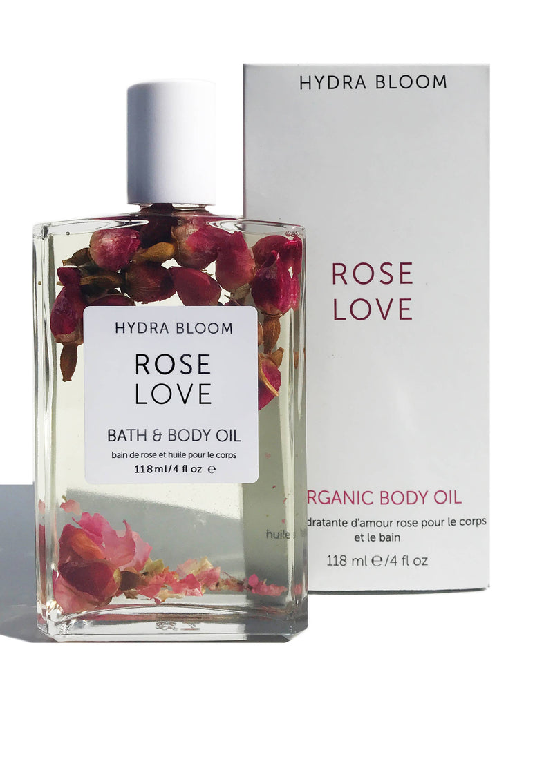 A clear glass bottle labeled "Hydra Bloom Beauty Rose Love Bath and Body Oil" filled with bath oil and dried rose petals, next to its packaging box on a white background.