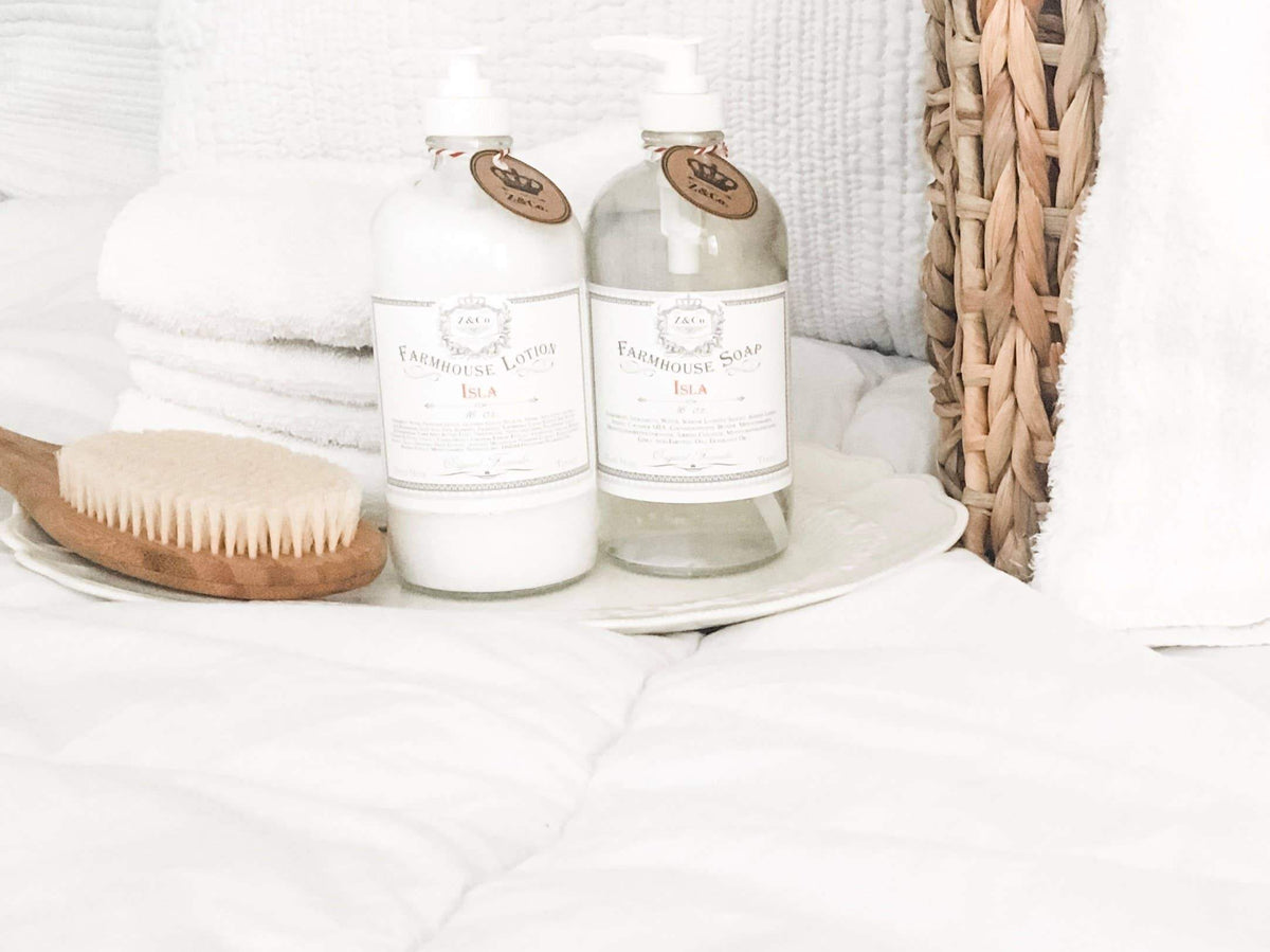 A tranquil bathroom setting featuring a bottle of Z&Co. Garden Gate Farmhouse Lotion and farmhouse soap infused with moisturizing natural ingredients, placed on a folded white towel, alongside a wooden brush and a woven basket.