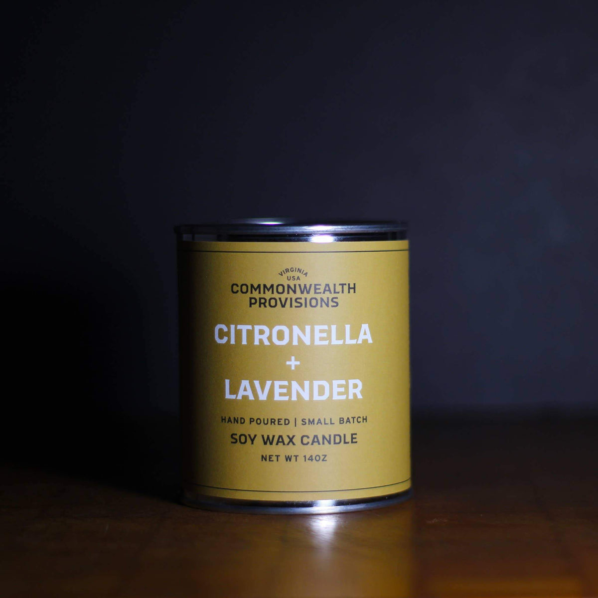 A soy wax Commonwealth Provisions Citronella + Lavender Candle in a metal tin on a dark, wooden surface with a soft focus background.