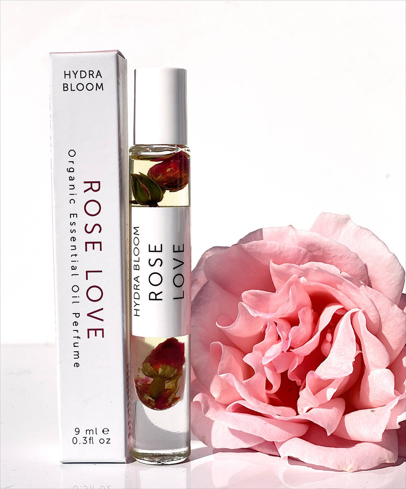 A bottle of Hydra Bloom Beauty Rose Love Essential Perfume Roll-on Oil with visible rose petals inside it, placed next to its packaging box and a vibrant pink rose on a white background.