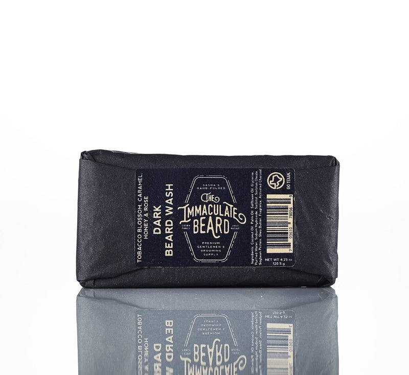 A black package of "The Immaculate Beard - Beard Wash Bar" with activated charcoal on a reflective surface, showcasing the front label with a vintage design.