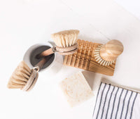A flat lay of eco-friendly cleaning supplies including a wooden saucepan brush, Z&Co. Lemon Farmhouse Solid-Block Dish Soap, a small bucket with soapy water, a bamboo soap dish, and a striped cloth on
