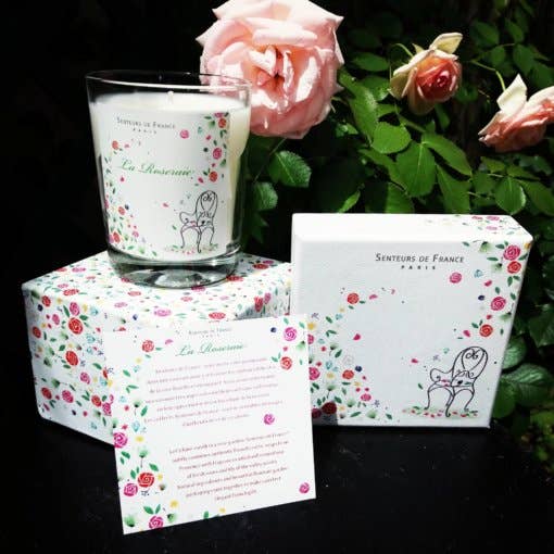 A Senteurs De France Candle Versailles Rose Garden in a glass container with floral prints, placed next to a similarly designed box and a small card, all set against a backdrop of pink roses in a dimly lit setting.